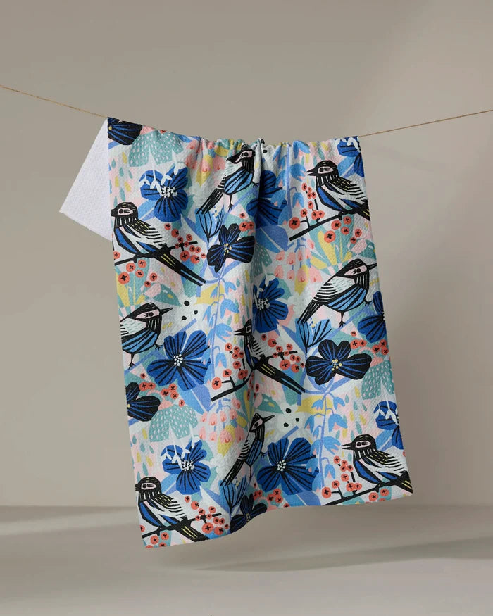 The "Birds And Berries" tea towel by Masha Volnova, made from recycled materials, displays black and white birds, blue flowers, and red berries on a neutral background.