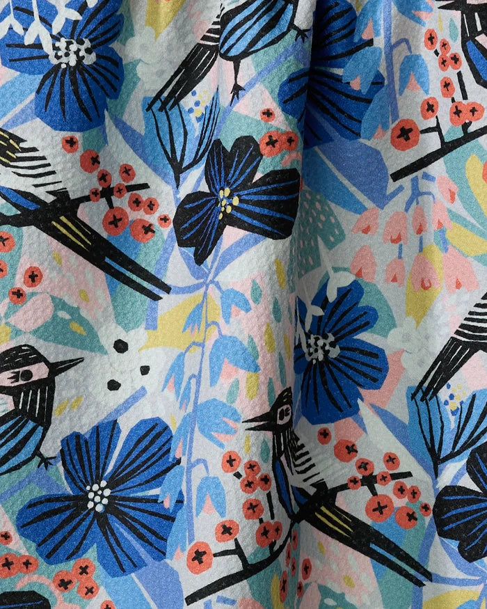 The "Birds And Berries" tea towel by Masha Volnova is made from recycled materials and features a colorful design with blue flowers, red berries, and stylized birds with black and white wings among vibrant abstract shapes.