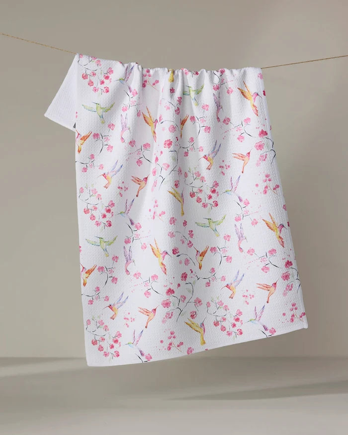 The Elena Fay "Birds and Blooms" tea towel is white, quick-drying, and textured. Hanging on a clothesline, it showcases colorful birds and pink cherry blossoms on a cheerful, delicate background that's plain and neutral.