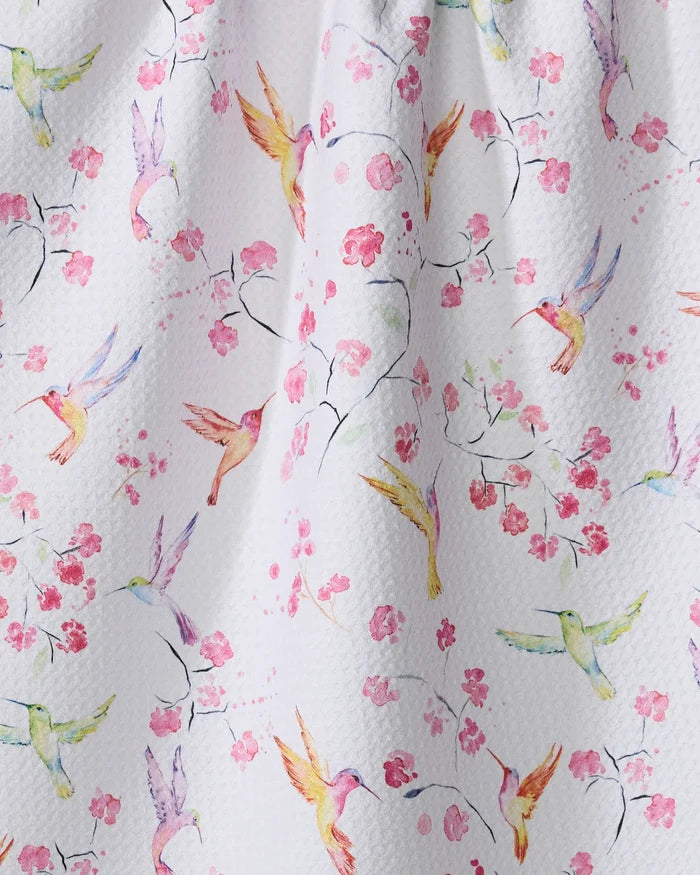 The "Birds and Blooms" tea towel by Elena Fay features a watercolor print of hummingbirds and cherry blossoms on white textured fabric, made from recycled materials.