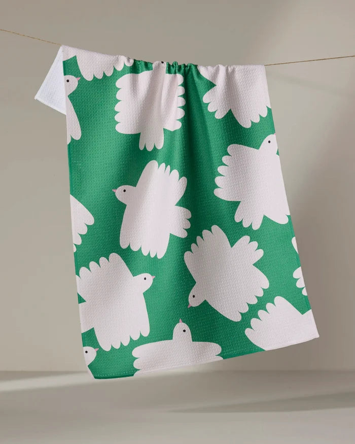 The "Birds" kitchen towel by Jennifer Bouron, made from recycled materials, features a green background with white stylized birds and hangs on a line against a neutral backdrop.