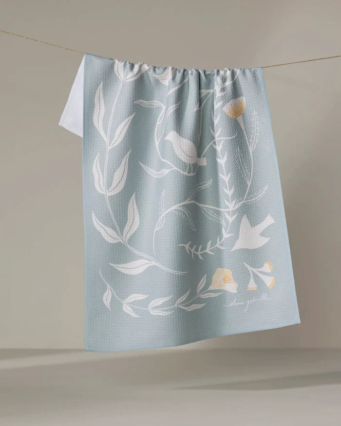 Birdsong by Elana Gabrielle is an eco-friendly, light blue kitchen towel featuring white botanical and bird illustrations with pale yellow accents, shown draped over a clothesline against a neutral backdrop.