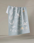 Birdsong by Elana Gabrielle is an eco-friendly, light blue kitchen towel featuring white botanical and bird illustrations with pale yellow accents, shown draped over a clothesline against a neutral backdrop.