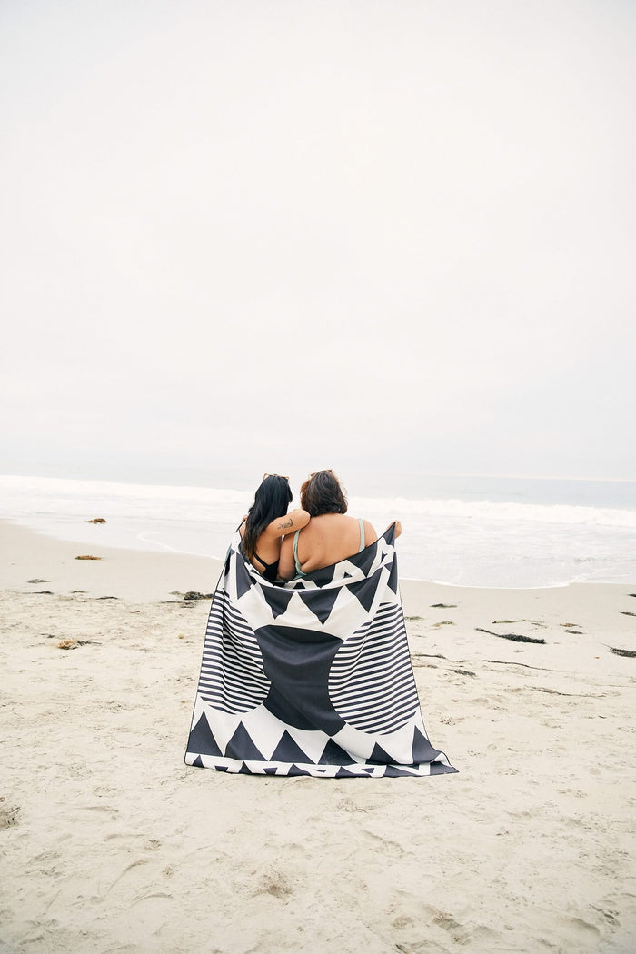 Two people sit side by side on a sandy beach, wrapped together in the GEOMETRY Black Stripes recycled beach blanket. The black and white geometric pattern stands out as they stay cozy by the shore, facing the ocean under an overcast sky.