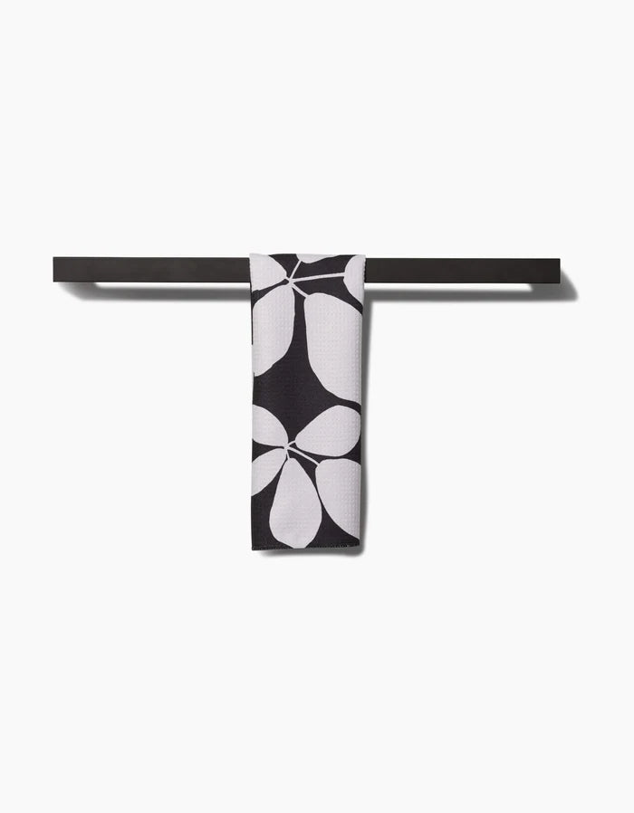 A black Blackberry bar towel by PrintedPlant, made from recycled materials, hangs centered on a black bar and shows a bold white petal or leaf pattern, all set against a plain white background.