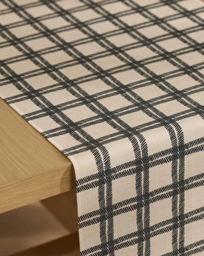 The Geometry Blackstone table runner, made from recycled fabric, features a beige base with a textured finish and a black plaid pattern of evenly spaced horizontal and vertical lines draped over a light wooden table.