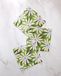 Three Bliss and Bloom cloths by GEOMETRY, featuring white daisies and green leaves on recycled fabric, are displayed on a white marble surface.
