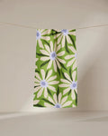 The GEOMETRY Bliss and Bloom green recycled kitchen towel, featuring a striking white daisy with a blue center, hangs on a clothesline against a neutral background.