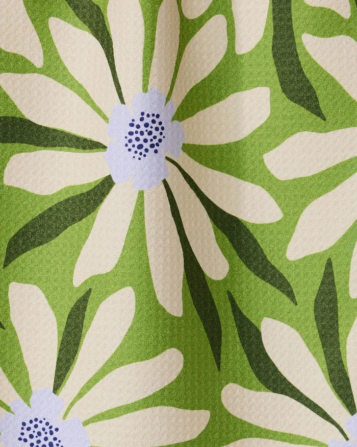 A close-up of the GEOMETRY Bliss and Bloom bar towel shows its ultra-absorbent, textured fabric featuring large white daisies with purple centers and green leaves on a light green background, displayed in soft folds.