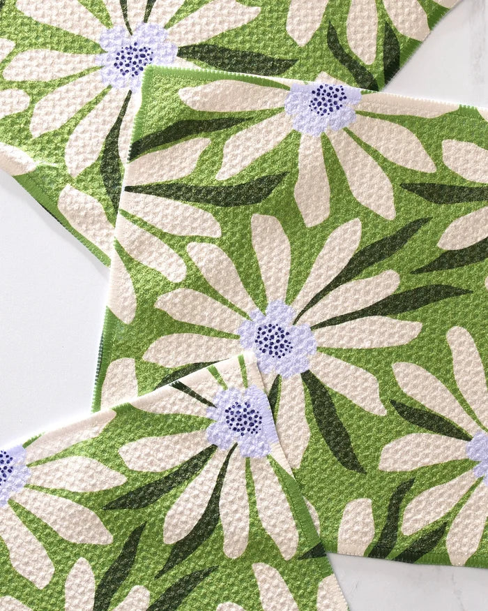 Bliss and Bloom dishcloths by GEOMETRY feature a white daisy with a purple center, large petals, and dark green leaves on green, overlapping each other. Made from recycled materials, these super-absorbent cloths brighten any space.
