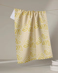 The Block Print Tulips tea towel by Lizzie Clark, crafted from recycled materials in a beige with yellow and white tulip pattern, hangs on a string. A white towel and stacked plates appear softly in the neutral background.
