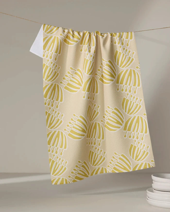 The Block Print Tulips tea towel by Lizzie Clark, crafted from recycled materials in a beige with yellow and white tulip pattern, hangs on a string. A white towel and stacked plates appear softly in the neutral background.