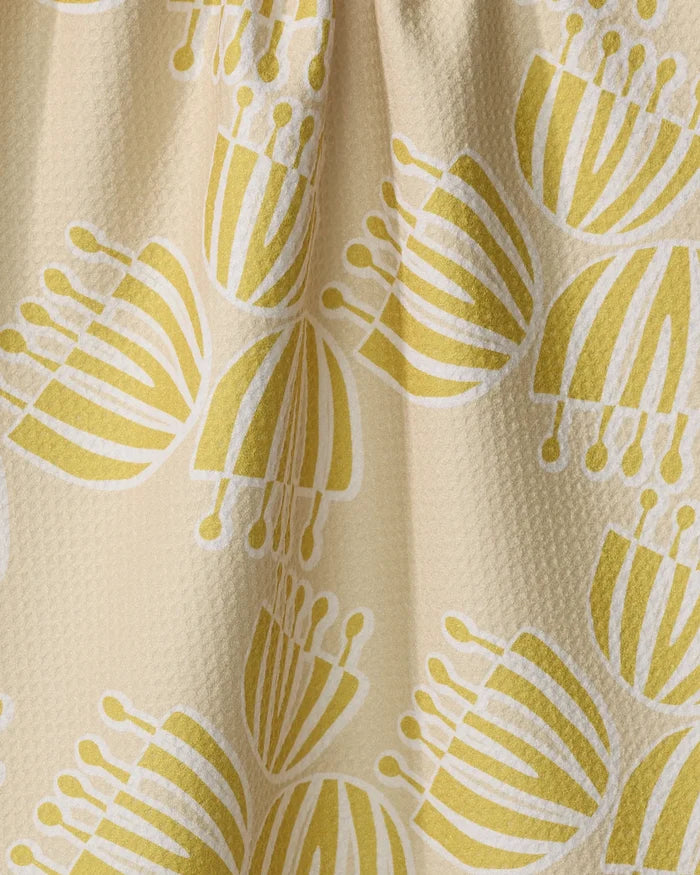 A close-up of the Lizzie Clark Block Print Tulips tea towel, crafted from recycled materials, shows a beige textured surface with a yellow and white abstract tulip pattern of clustered, striped petal shapes with circular ends.