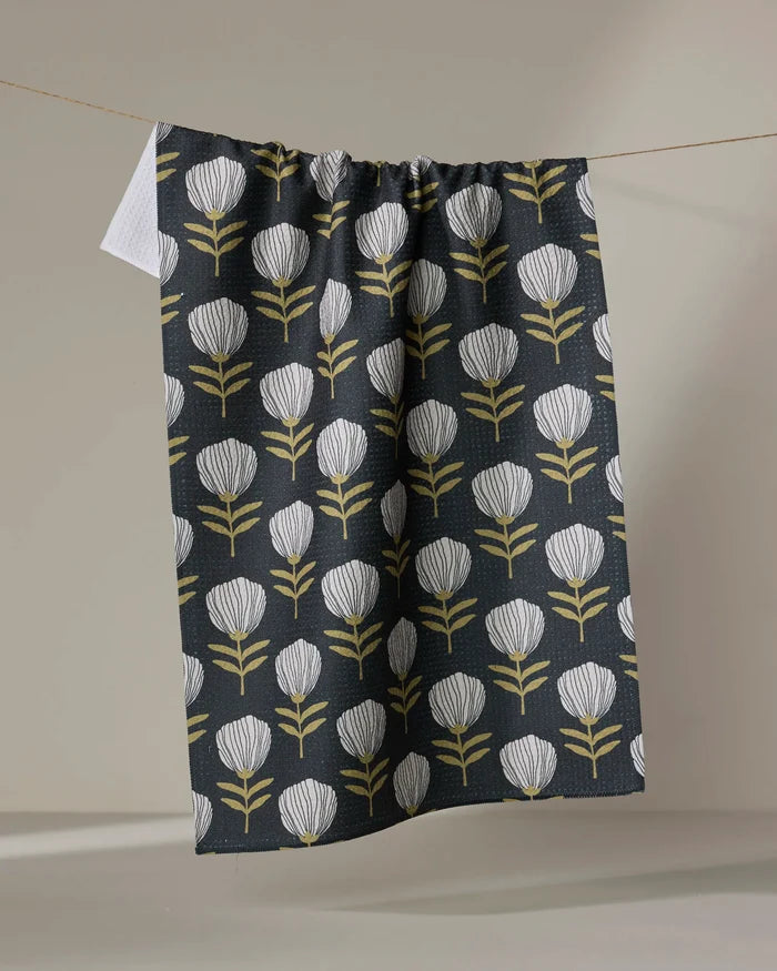The Anna Christian Blomma tea towel, featuring a dark background and a repeating pattern of white and yellow stylized flowers, hangs on a clothesline against a plain backdrop.