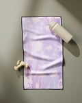 The GEOMETRY Bloom Blend ultra-absorbent fitness towel with a floral pattern rests on a gray surface beside a beige water bottle and two small, light-colored dumbbells.