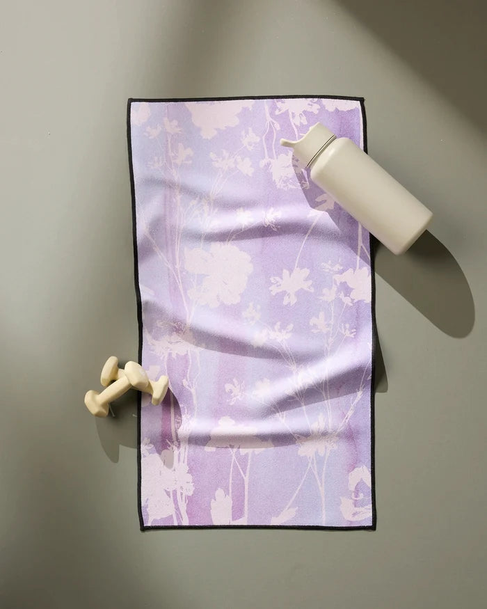 The GEOMETRY Bloom Blend ultra-absorbent fitness towel with a floral pattern rests on a gray surface beside a beige water bottle and two small, light-colored dumbbells.
