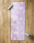 The GEOMETRY Bloom Blend yoga mat with white floral patterns is on a wooden floor, topped with a silicone grip yoga towel. A water bottle sits at the top left and gray yoga blocks are at the bottom right of the mat.