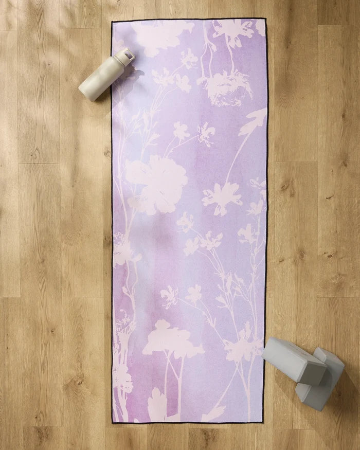 The GEOMETRY Bloom Blend yoga mat with white floral patterns is on a wooden floor, topped with a silicone grip yoga towel. A water bottle sits at the top left and gray yoga blocks are at the bottom right of the mat.