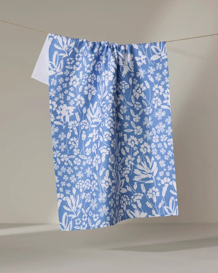 The Bloom Me Over Floral dish towel by Hufton Studio, featuring a blue and white floral pattern, hangs absorbently on a clothesline against a simple light backdrop.