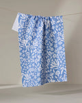 The Bloom Me Over Floral dish towel by Hufton Studio, featuring a blue and white floral pattern, hangs absorbently on a clothesline against a simple light backdrop.