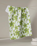 The Lizzie Clark "Blooming Blossoms" kitchen towel, featuring a green floral and leaf design on white, hangs above a stack of white plates against a neutral background.