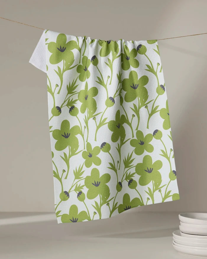 The Lizzie Clark "Blooming Blossoms" kitchen towel, featuring a green floral and leaf design on white, hangs above a stack of white plates against a neutral background.