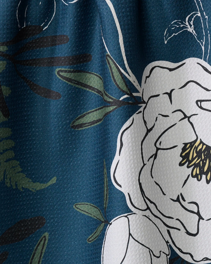 A close-up of the Blooming Possibilities tea towel by Ashley Celena, showing its blue Geoweave fabric with a textured surface, white floral pattern, and green leafy accents outlined in black.