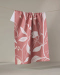 The Elana Gabrielle Blooms recycled kitchen towel, featuring a pink and white botanical leaf pattern, hangs neatly on a clothesline against a plain, neutral background.
