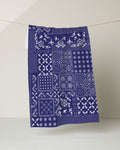 The Geometry Blossoming Bandanna Blue kitchen tea towel, crafted from recycled materials, features white floral and geometric patchwork on dark blue and hangs against a light background.