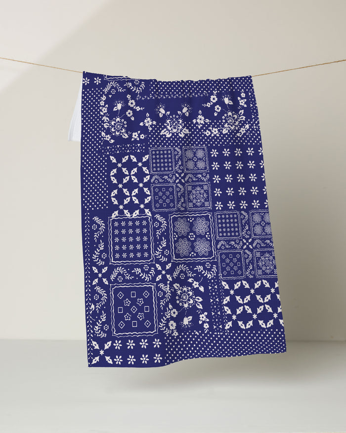 The Geometry Blossoming Bandanna Blue kitchen tea towel, crafted from recycled materials, features white floral and geometric patchwork on dark blue and hangs against a light background.