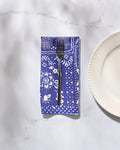 A fork rests on Geometry’s Blossoming Bandanna Blue reusable cloth napkin, featuring white floral patterns, next to a scalloped white plate on a marble surface.