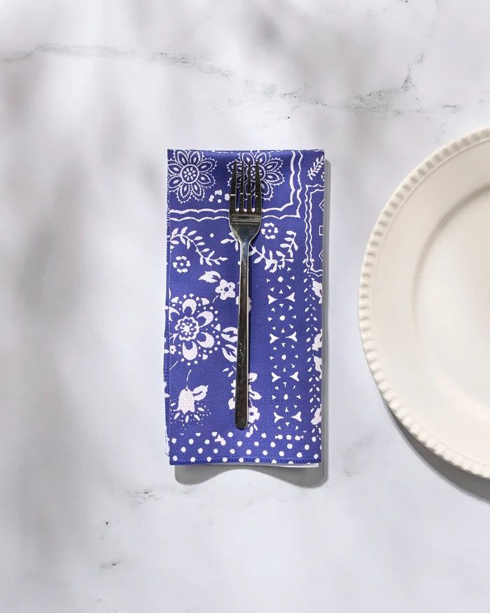 A fork rests on Geometry’s Blossoming Bandanna Blue reusable cloth napkin, featuring white floral patterns, next to a scalloped white plate on a marble surface.