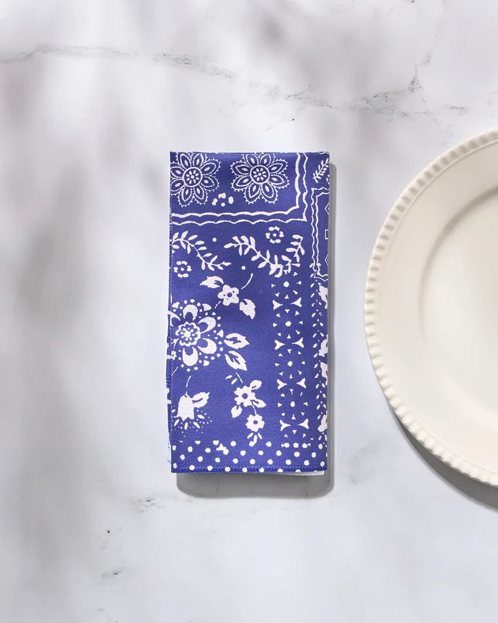 The Geometry "Blossoming Bandanna Blue" reusable cloth napkin, featuring white floral and geometric patterns, is neatly folded beside a white ribbed-edge plate on light marble, with soft shadows adding elegance to the scene.