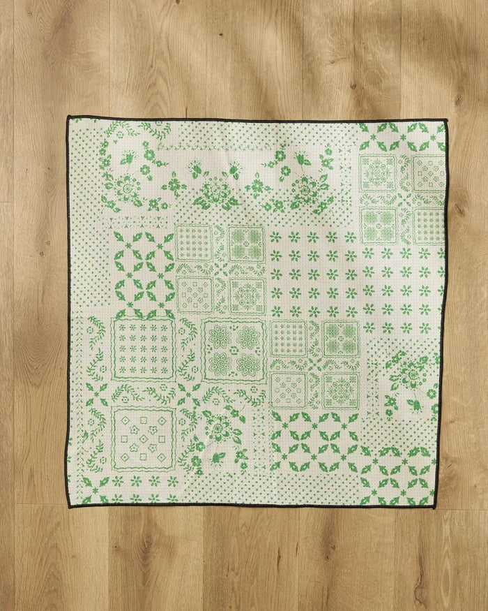 The Blossoming Bandanna Green quilt by Geometry, made from recycled materials, features green floral and geometric patterns on a white background with a black border, and is displayed flat on a light wooden floor.