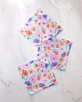The GEOMETRY Blossoming Love set features three square dishcloths with colorful orange, purple, pink, and beige floral patterns with green leaves, arranged on a marble-like surface. Made from recycled materials for vibrant style.
