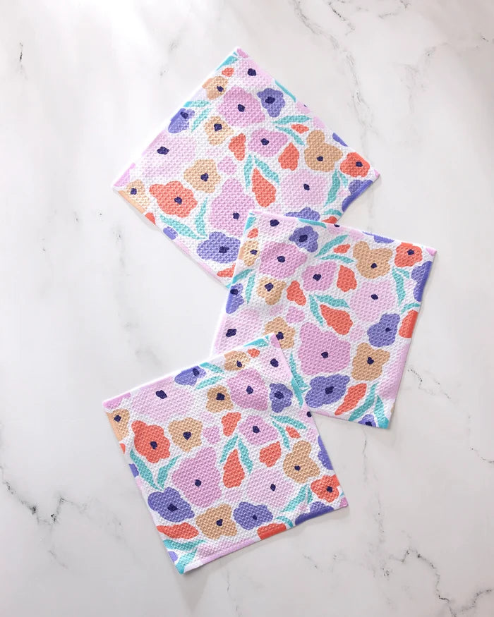 The GEOMETRY Blossoming Love set features three square dishcloths with colorful orange, purple, pink, and beige floral patterns with green leaves, arranged on a marble-like surface. Made from recycled materials for vibrant style.