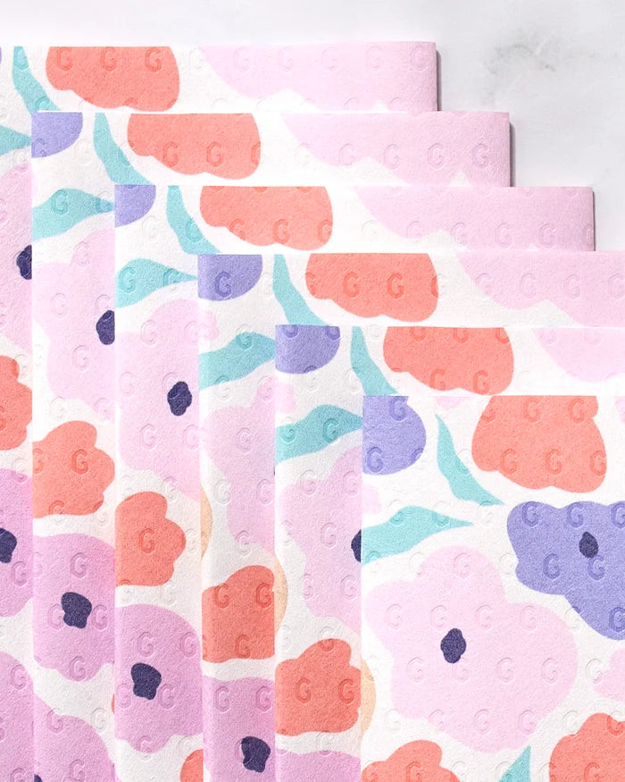 Blossoming Love reusable towels by GEOMETRY, with pastel floral patterns in pink, orange, purple, and blue, are fanned out on a light surface. Featuring a subtle embossed G design, they’re an eco-friendly alternative to paper towels.