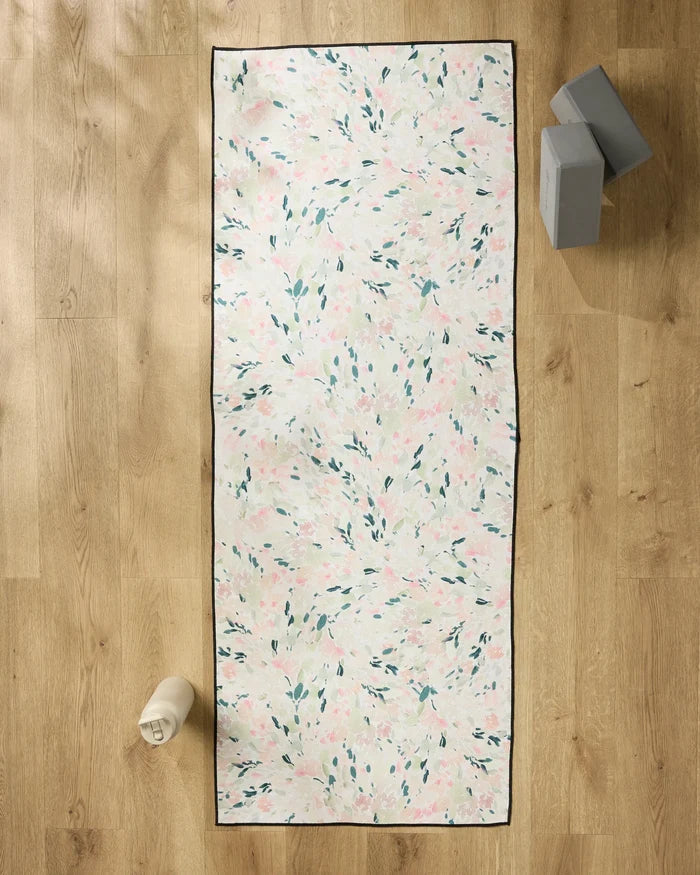 The Blossoms yoga mat by Anee Shah, featuring green, pink, and white florals, rests on a wooden floor with a white water bottle, two gray blocks stacked nearby, and a soft non-slip yoga towel for extra comfort.