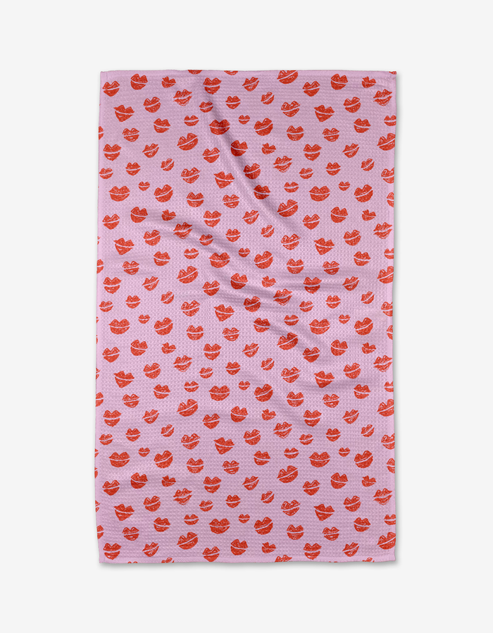 The Kayla Ann Blowing Kisses tea towel is pink and rectangular, featuring a red lips pattern. Made from recycled materials, it’s shown laid flat with slight wrinkles visible.