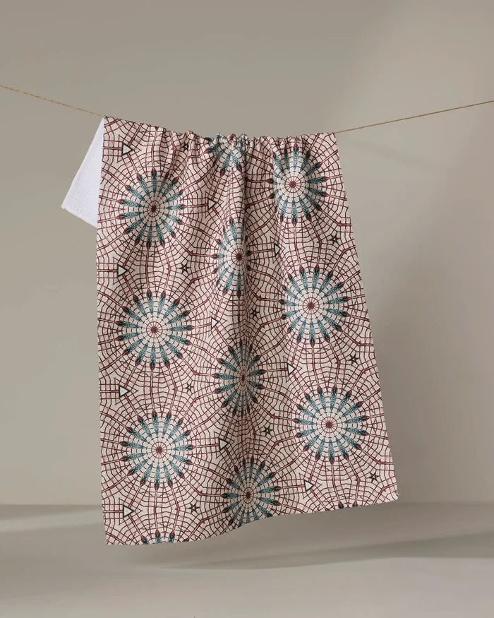 The Katherine Berkeley Blue Aster Foxtrot kitchen towel, featuring blue and red geometric starburst patterns and made from recycled materials, hangs on a clothesline against a plain, neutral background.