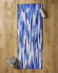 The GEOMETRY Blue Blast yoga mat is laid out on a wooden floor, accompanied by a silicone grip yoga towel, two gray yoga blocks, and a white water bottle.