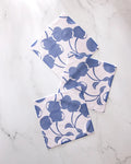 Three Geometry "Blue Cherry Dreams" napkins with a blue floral pattern are arranged on a white marble surface, slightly overlapping at different angles.