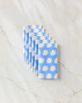 Six folded Megan Roy Blue Daisies reusable towels, featuring a white and yellow daisy pattern, are neatly fan-arranged on a white marble surface with light brown veins.