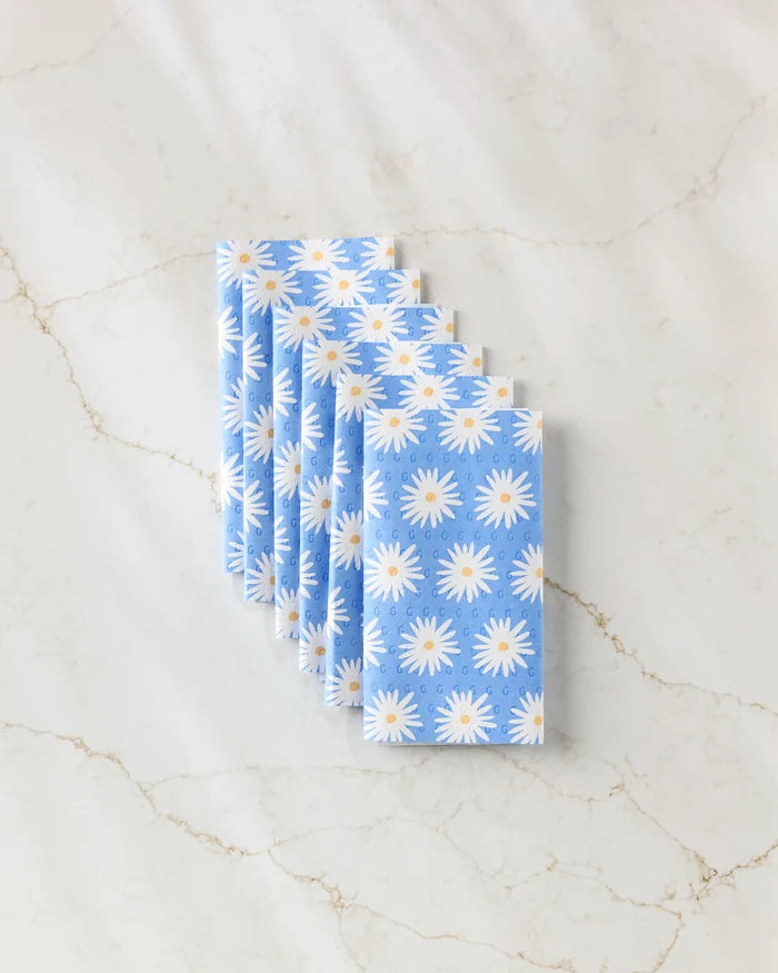Six folded Megan Roy Blue Daisies reusable towels, featuring a white and yellow daisy pattern, are neatly fan-arranged on a white marble surface with light brown veins.