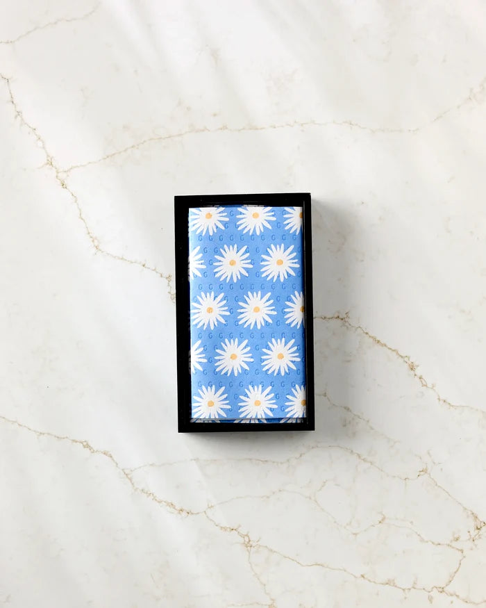 A rectangular bar of Megan Roy's Blue Daisies is wrapped in blue, eco-friendly paper towels with a white daisy pattern and placed in a black tray on a light marble-patterned surface.