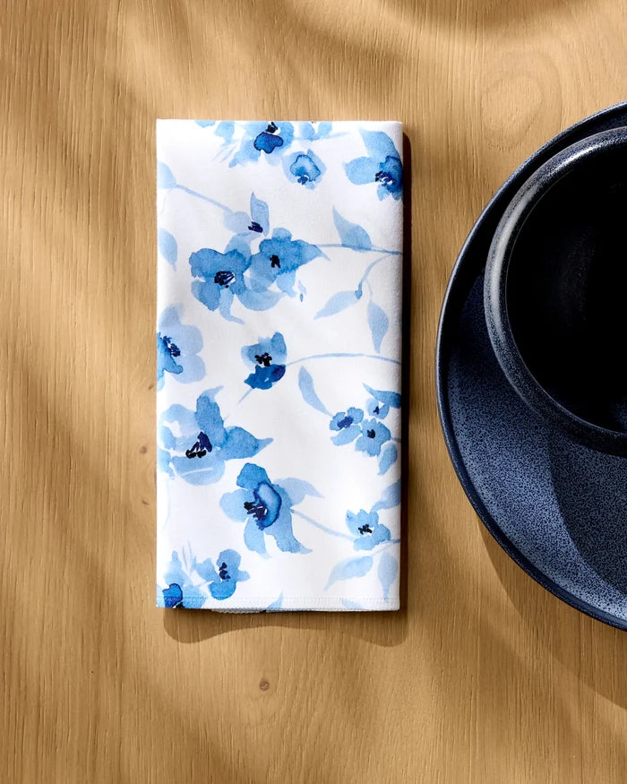 The Aubrey Fairchild Blue Floral napkin, a folded white absorbent dinner napkin with blue floral patterns, rests on a light wooden table beside a dark blue plate and cup.