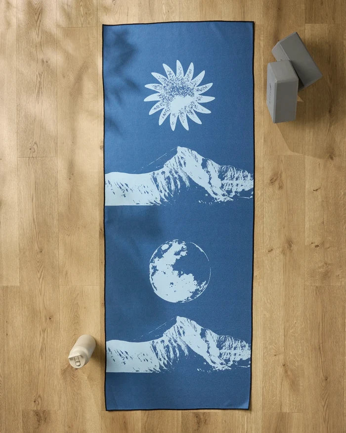 The Blue Moon yoga mat by Graphics and Grain features white mountain, sun, and moon designs. It’s shown on a wooden floor with two gray yoga blocks, a white water bottle, and a non-slip towel for your practice.