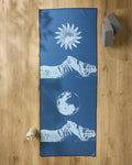 The Blue Moon yoga mat by Graphics and Grain features white mountain, sun, and moon designs. It’s shown on a wooden floor with two gray yoga blocks, a white water bottle, and a non-slip towel for your practice.