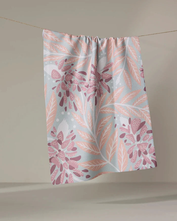 The Bluebonnet tea towel by Julianne Haness, featuring a pink and peach leaf and floral pattern on pastel colors, made from recycled materials, hangs on a clothesline against a neutral background.