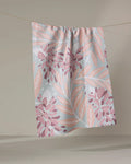 The Bluebonnet tea towel by Julianne Haness, featuring a pink and peach leaf and floral pattern on pastel colors, made from recycled materials, hangs on a clothesline against a neutral background.